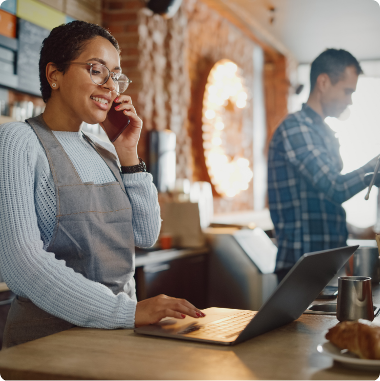 Woman wearing apron talking on the phone and on her laptop. 